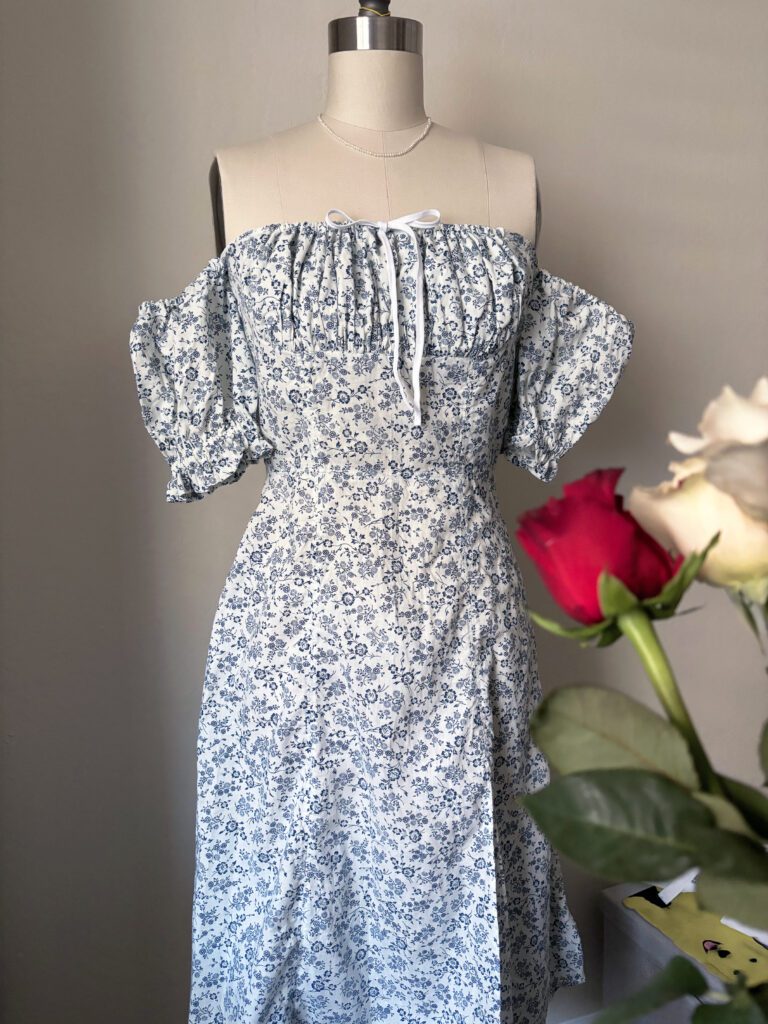 A full view of the modified Anthea Milkmaid Dress in blue floral print, showcasing the ruched bust, a bow knot, puffy sleeves and a leg split.